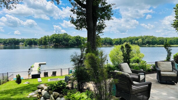 A patio with wicker furniture overlooking a lake, surrounded by greenery and a stone wall. - Always East Lake-  all sports lake, on Little Hart Lake, swim, kayak, fishing (Twin Lake)