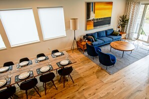 Un salon moderne avec un canapé bleu, une table à manger dressée pour six personnes et une grande fenêtre équipée de stores.
