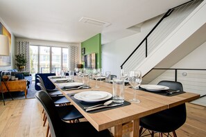 Un coin repas est aménagé avec une longue table en bois, dressée pour six personnes, et entourée de chaises noires. Un escalier est visible sur la droite, menant à un niveau supérieur.