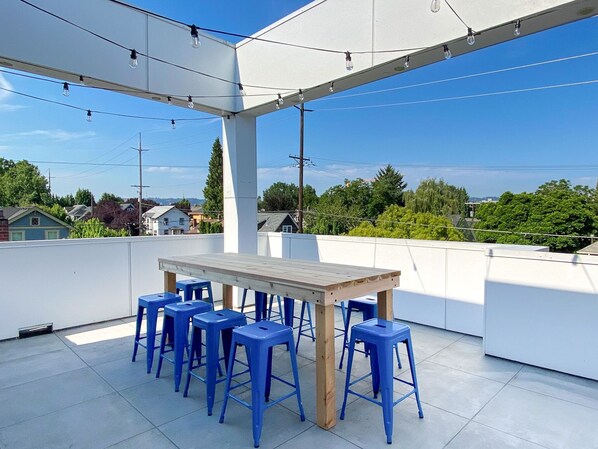 Une terrasse extérieure avec une table en bois et des tabourets bleus, entourée de murs blancs et illuminée de guirlandes lumineuses.