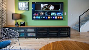Un salón moderno con un televisor de pantalla plana montado en una pared verde, un banco de madera, una silla metálica y un tapete estampado.