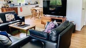 Een moderne woonkamer met een zwarte leren bank, een bijzettafeltje en een televisie. Achtergrond is een keuken met witte kasten en een raam.