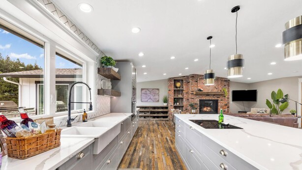 A modern kitchen with a large island, white cabinetry, and a brick accent wall. - 3 Acres Remodeled Home With Hot Tub Walking Distance to Beach! Garden & Fire Pit (Coeur d Alene)