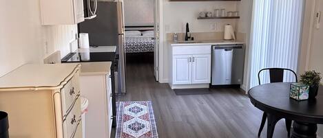 Cocina compacta con armarios blancos, microondas y refrigerador. Cuenta con un comedor con mesa y sillas, y una alfombra pequeña en el suelo.
