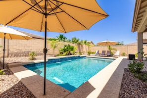 A swimming pool with a tiled edge, surrounded by a pebble border and a low wall. There are lounge chairs and a large umbrella providing shade. - Relaxing stay with FREE heated pool! The Rimrock Retreat (Surprise)