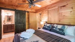 Een slaapkamer in een houten blokhut met een bed, nachtkastje, lamp, plafondventilator en een houten deur.