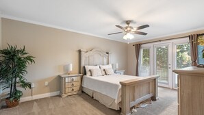 Quarto com cama, mesas de cabeceira, ventilador de teto, planta em vaso e vista através de uma porta de vidro.