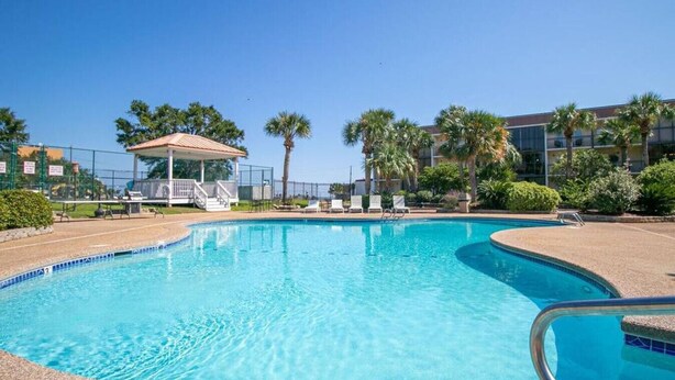 Piscina cristalina com um gazebo, palmeiras e um edifício ao fundo.