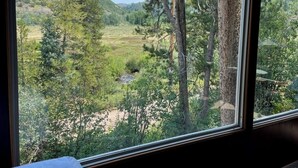 A room with a large window overlooking a forested area with a stream. - Cozy Mountain Paradise (Kamas)