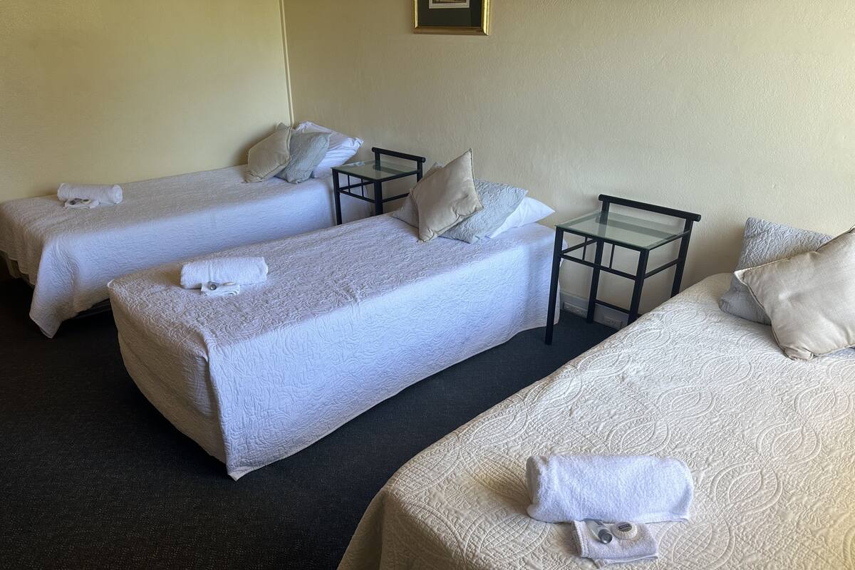 A hotel room with three single beds, each with a pillow and a rolled towel. There is a small table between the beds.