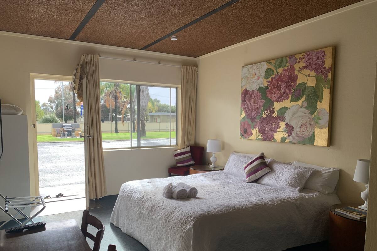 A hotel room with a large bed, a painting of flowers on the wall, a desk with a chair, and a view of the outside.