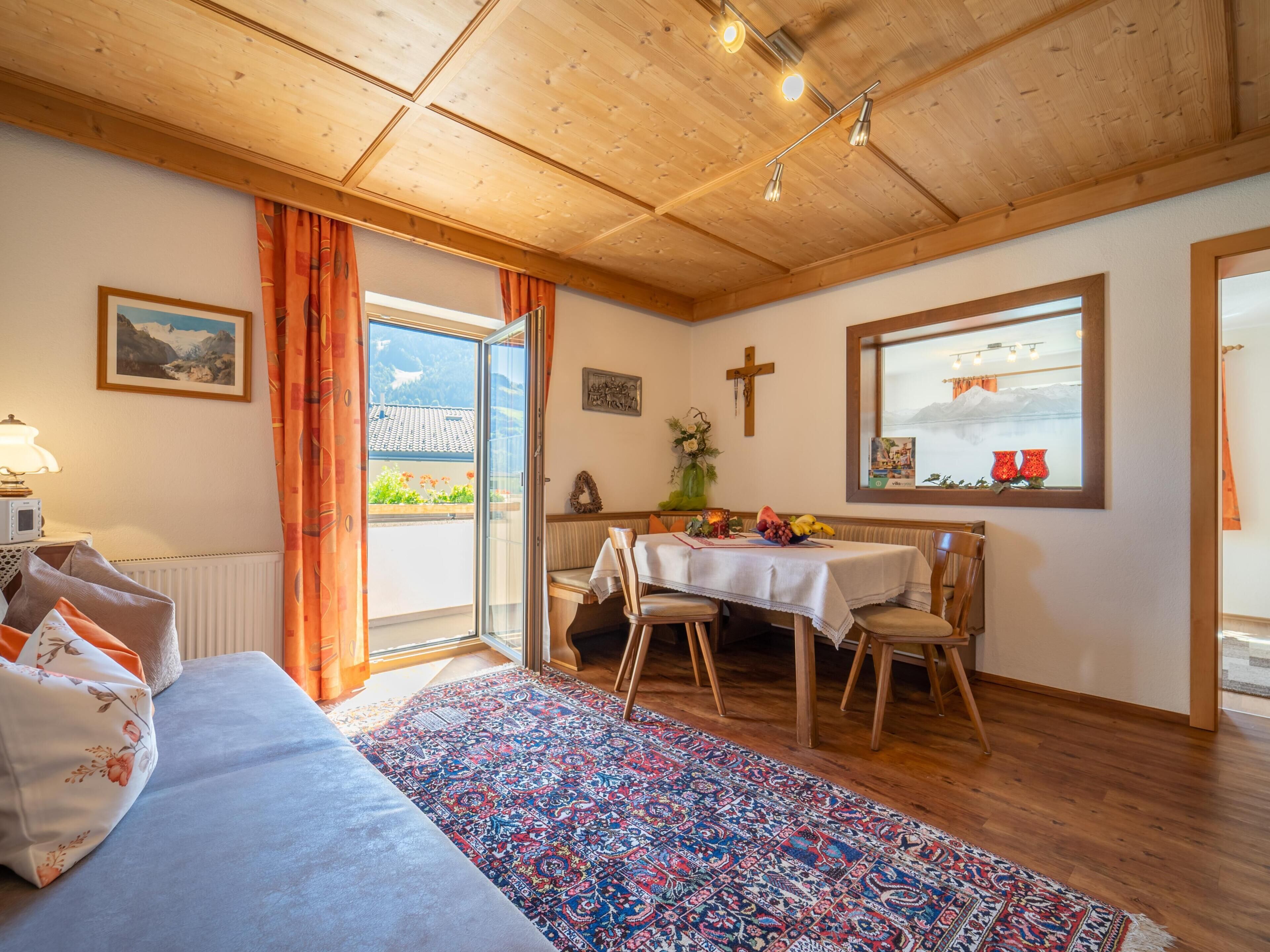 A cozy room with a bed, a dining table with chairs, a window with a view of mountains, and a wooden ceiling.