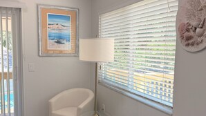 Ein Zimmer mit weißem Sessel, Stehlampe, gerahmtem Gemälde und einem Fenster mit Jalousien.