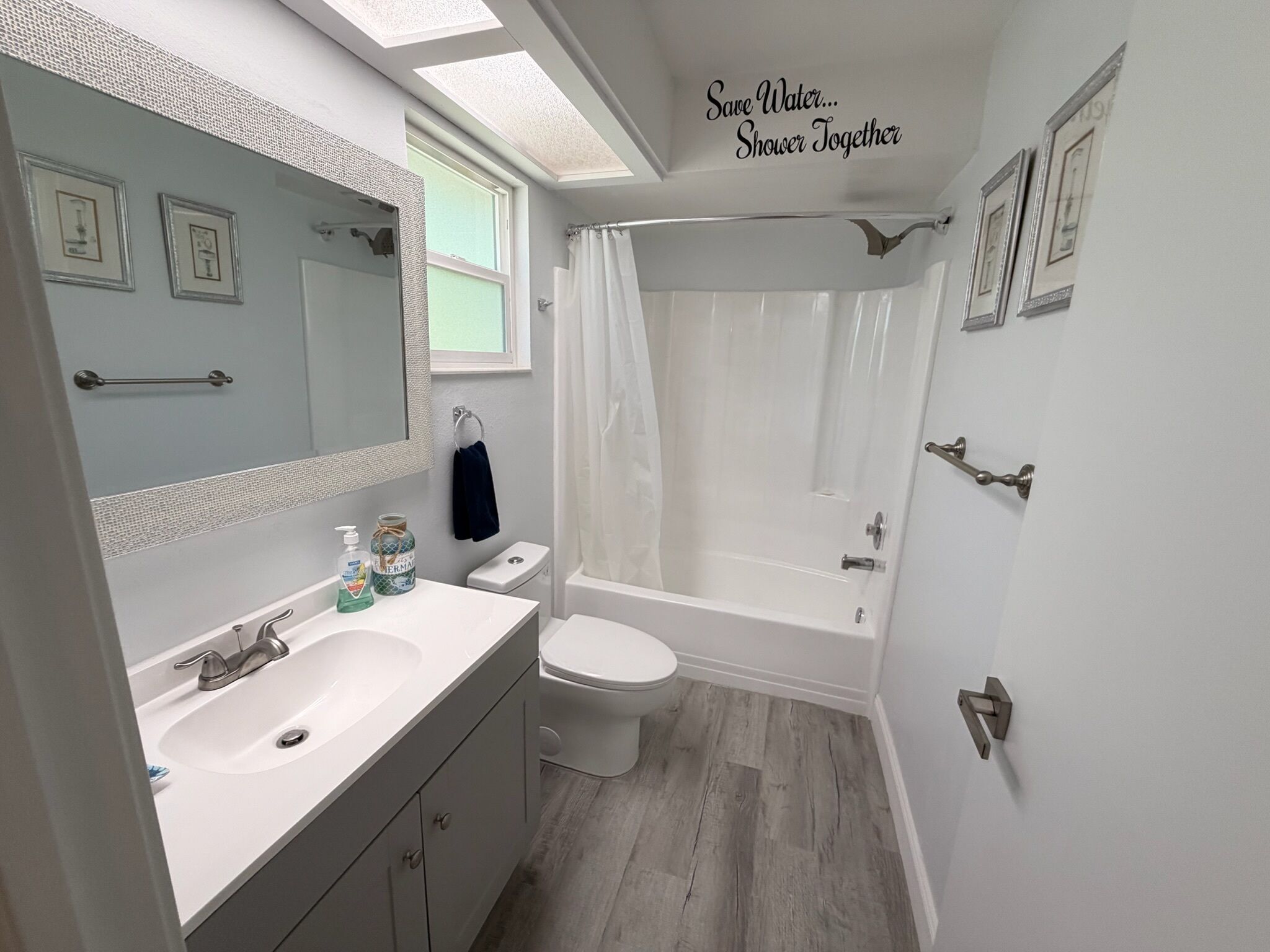 Une salle de bain équipée d’un lavabo, de toilettes et d’une baignoire, ainsi que d’un porte-serviettes et d’une pancarte indiquant : « Économisez l’eau… prenez votre douche à deux ».