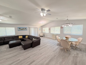 A spacious living area with a sectional sofa, a dining table with chairs, a ceiling fan, and large windows with blinds.