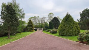 Een verharde oprit leidt naar een toegangshek, omgeven door keurig onderhouden gazons en diverse bomen.