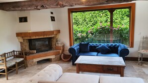 Een gezellige woonkamer met een stenen open haard, een blauwe bank en uitzicht op weelderige begroeiing door een groot raam.