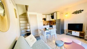 A modern living room with a sofa, a dining table, and a kitchen area with a TV and refrigerator. - Villa Grand Bleu Odalys (Vendres PLAGE)