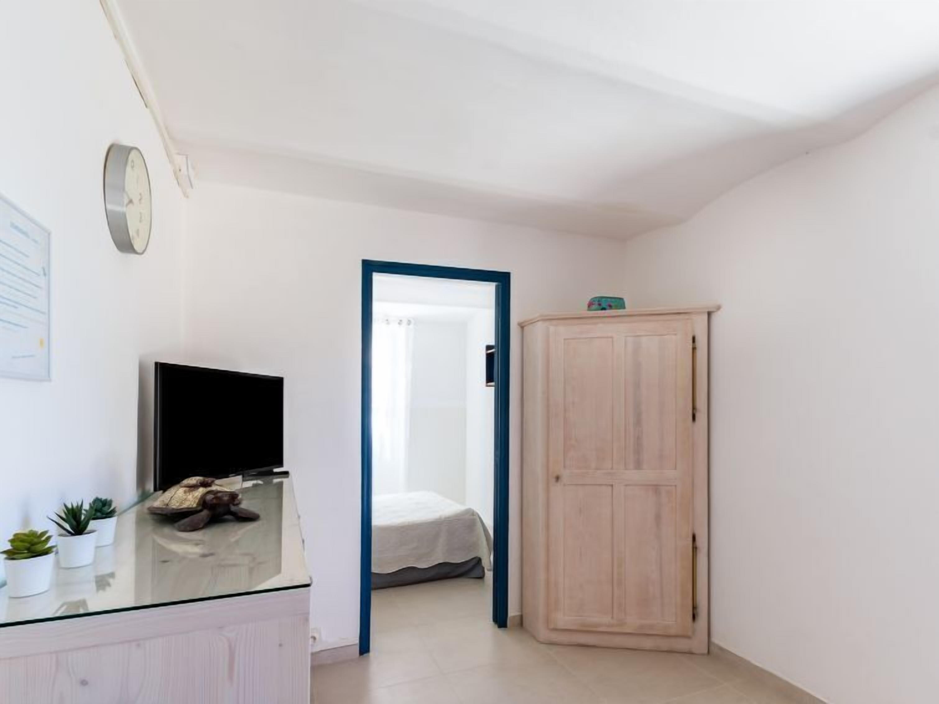 Une pièce avec un bureau à plateau en verre, un téléviseur à écran plat, une armoire en bois et un lit à l’arrière-plan.
