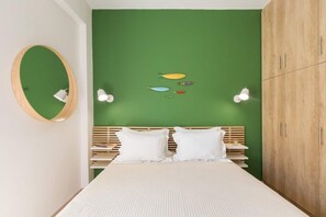 A bedroom with a green wall, a wooden wardrobe, a bed with white bedding, and a round mirror.