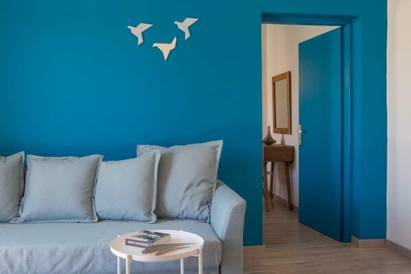 Une pièce avec un mur bleu, un canapé gris, une petite table ronde sur laquelle se trouve un livre, et une porte qui donne sur une autre pièce.