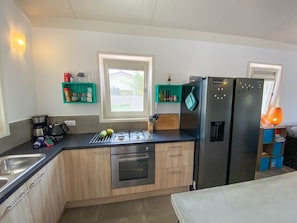 Eine kompakte Küche mit Gasherd, Backofen und einem schwarzen Kühlschrank. Zwei Fenster sorgen für natürliches Licht, und ein türkisfarbenes Regal beherbergt Küchenutensilien.