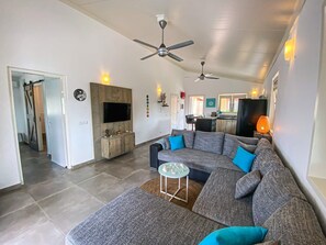 Ein modernes Wohnzimmer mit einem Ecksofa, einem Couchtisch und einem Fernseher. Es gibt Deckenventilatoren, eine Küche im Hintergrund und eine Tür zu einem weiteren Raum.