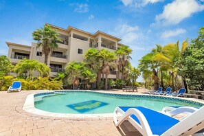 A swimming pool with lounge chairs, surrounded by palm trees and a multi-story building.