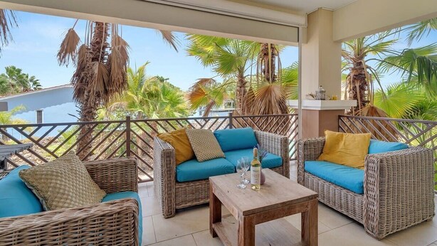 Un espacio de estar techado al aire libre con muebles de mimbre y una mesa de madera, con vistas a palmeras y un cielo despejado.