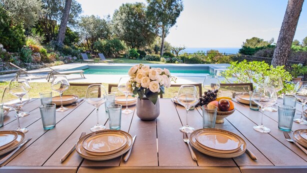An outdoor dining area with a pool view, neatly set tables with plates, glasses, and a vase of flowers.
