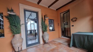 Une pièce avec un sol carrelé, deux grandes plantes en pot, une table recouverte d’une nappe verte et une porte donnant sur un balcon.