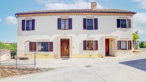 Une maison à deux étages, avec un toit en tuiles, de nombreuses fenêtres équipées de volets et une entrée en pierre.
