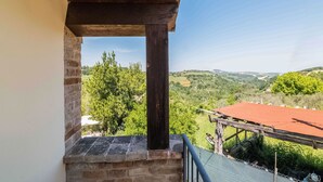 A balcony with a stone wall, wooden railing, and a view of a green landscape with trees and hills.
