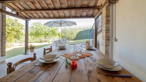 An outdoor dining area with a wooden table set for four, surrounded by chairs, under a wooden pergola. - Holiday home in Torchiaro with garden (Torchiaro)