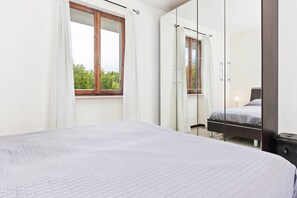 A bedroom with a large bed, a window with curtains, a mirror, and a view of greenery outside.