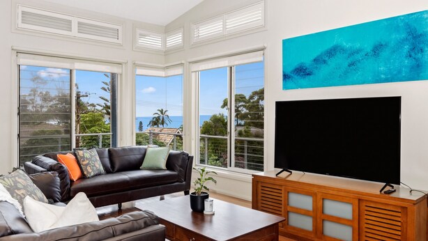 A living room with a leather sofa, a coffee table, a TV, and large windows with a view of the ocean. - Wallace Waves in Mollymook (Mollymook)