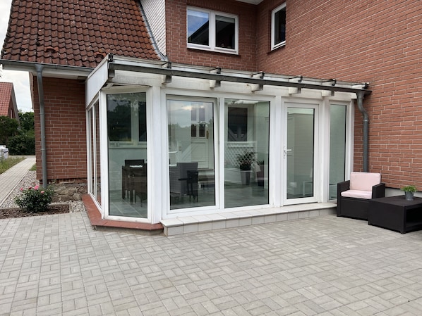 Un patio cubierto con cerramiento de vidrio, una casa de ladrillo y mobiliario exterior.