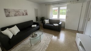 Ein modernes Wohnzimmer mit zwei dunklen Sofas, einem Glastisch und einem weißen Teppich. Ein großes Fenster mit Vorhängen, ein Blumentopf und eine Tür mit Heizkörper.