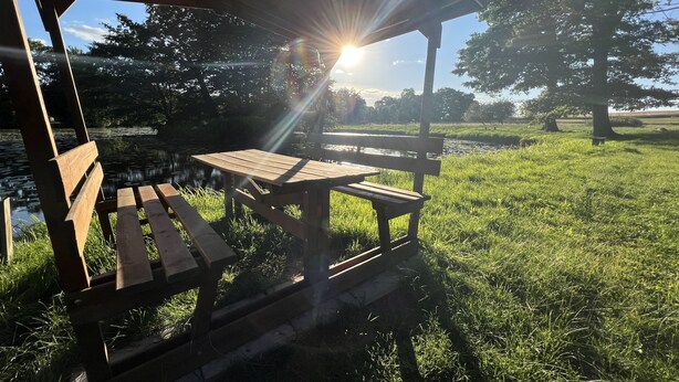 Ein hölzernes Pavillon mit Bänken und einem Picknicktisch, umgeben von Gras und Bäumen, wobei die Sonne durch das Dach scheint.