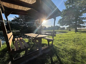 Un pabellón de madera con bancos y una mesa de picnic, rodeado de césped y árboles, con el sol brillando a través del techo.