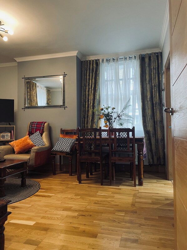 A cozy living room with a wooden floor, a sofa, a dining table with chairs, and a large window with curtains. - Cozy, quiet flat near Thames River, shops, restaurant & ublic transport (London)