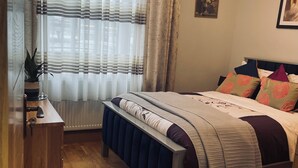 Ein Schlafzimmer mit einem Bett, einer Sitzbank, einem Schreibtisch, einer Topfpflanze und einem Fenster mit Vorhängen.