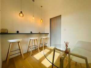 A modern kitchen with a glass dining table, wooden stools, and pendant lighting.