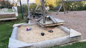 Un área de juegos con arena y estructuras de madera, incluyendo columpios y una mesa de picnic.