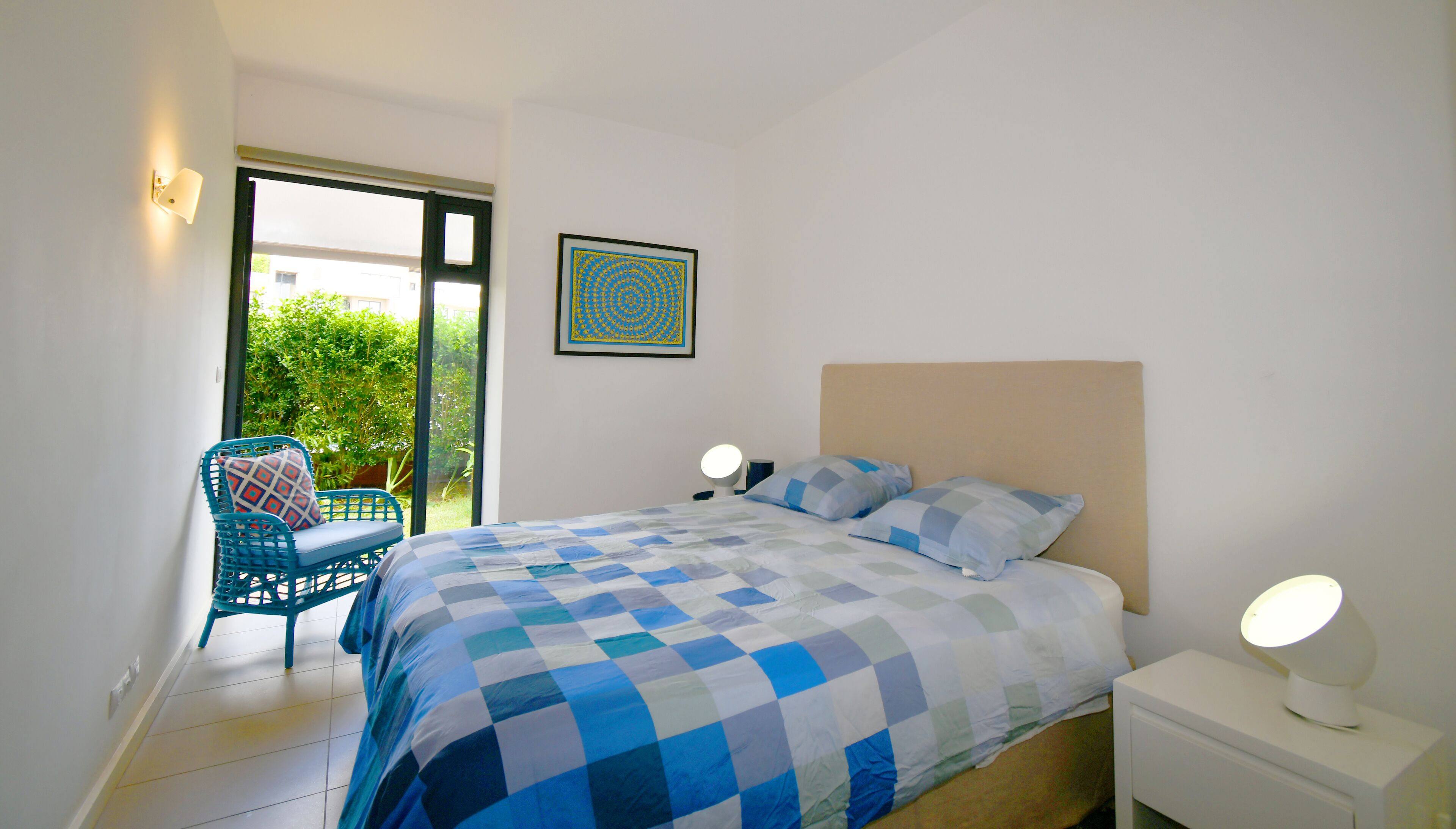 Une chambre avec une couvre-lit à carreaux, une chaise, une table de chevet avec une lampe et une fenêtre donnant sur un paysage verdoyant.