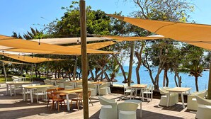 Un espace extérieur aménagé avec des tables et des chaises, protégé du soleil par de grands parasols, et offrant une vue sur une étendue d’eau.