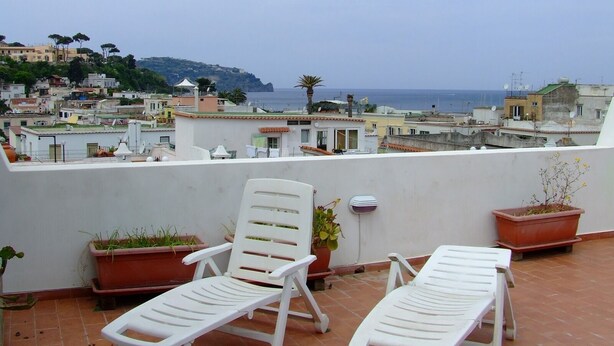 Eine Terrasse mit zwei weißen Liegestühlen, Kübelpflanzen und Blick auf das Meer und die Bebauung.