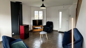 Una sala de estar con televisor, un mueble rojo, una silla azul, un taburete blanco y una lámpara de techo negra.