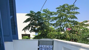 Un balcon avec une table et des chaises carrelées, un pot de fleurs aux fleurs rouges, et une vue sur la verdure et un bâtiment en arrière-plan.
