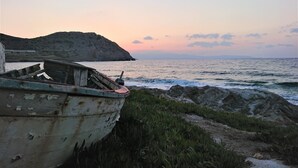 Un bateau amarré à un rivage rocheux, avec une chaîne de montagnes en arrière-plan et un coucher de soleil sur l’océan.